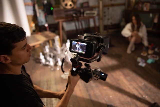 A person operates a professional video camera on a stabiliser, focusing on a woman sitting on the wooden floor in a dimly lit room with scattered objects and vintage decor. The camera screen displays the scene being filmed.